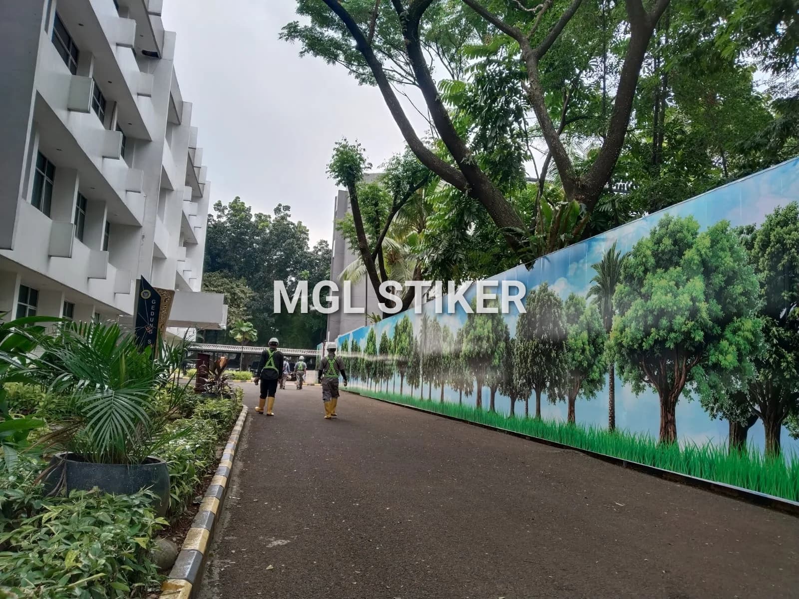 Pagar proyek panjang dilapisi stiker print digital dari MGL Stiker, menampilkan motif pepohonan hijau dan langit biru yang cerah, menciptakan suasana asri di area proyek. Terlihat beberapa pekerja berjalan di samping pagar.